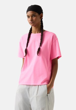 Unge med flettet hår iført en lyserød, oversized T-shirt og lysegrå bukser, stående med den ene hånd i lommen.