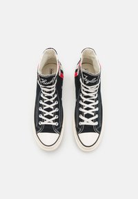 Sneakers alte in tela nera con lacci bianchi, punta in gomma, dettagli rossi e blu, e "Chuck Taylor" ricamato di lato.