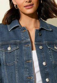 Giacca di denim blu scuro con colletto, bottoni argentati e due tasche sul petto. Il tessuto ha una texture classica con cuciture visibili.