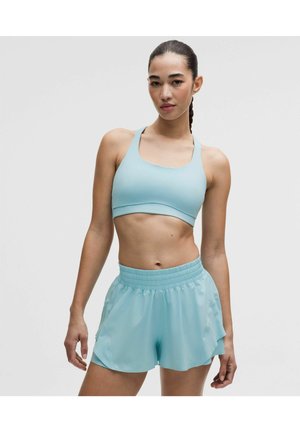 Fit kvinde med fletninger iført lyseblå sportstop og matchende atletikshorts, stående mod en ensfarvet grå baggrund.