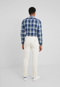 Pantalon blanc à coupe ajustée, associé à une chemise à carreaux bleus et verts. La chemise a des manches longues et des poignets boutonnés.