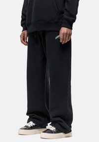 Zwarte sweatshirt en sweatpants gemaakt van katoenmix stof. De sweatpants hebben een klein wit logo, een relaxte pasvorm en een geborduurd zoom. Zwarte sneakers met witte veters.