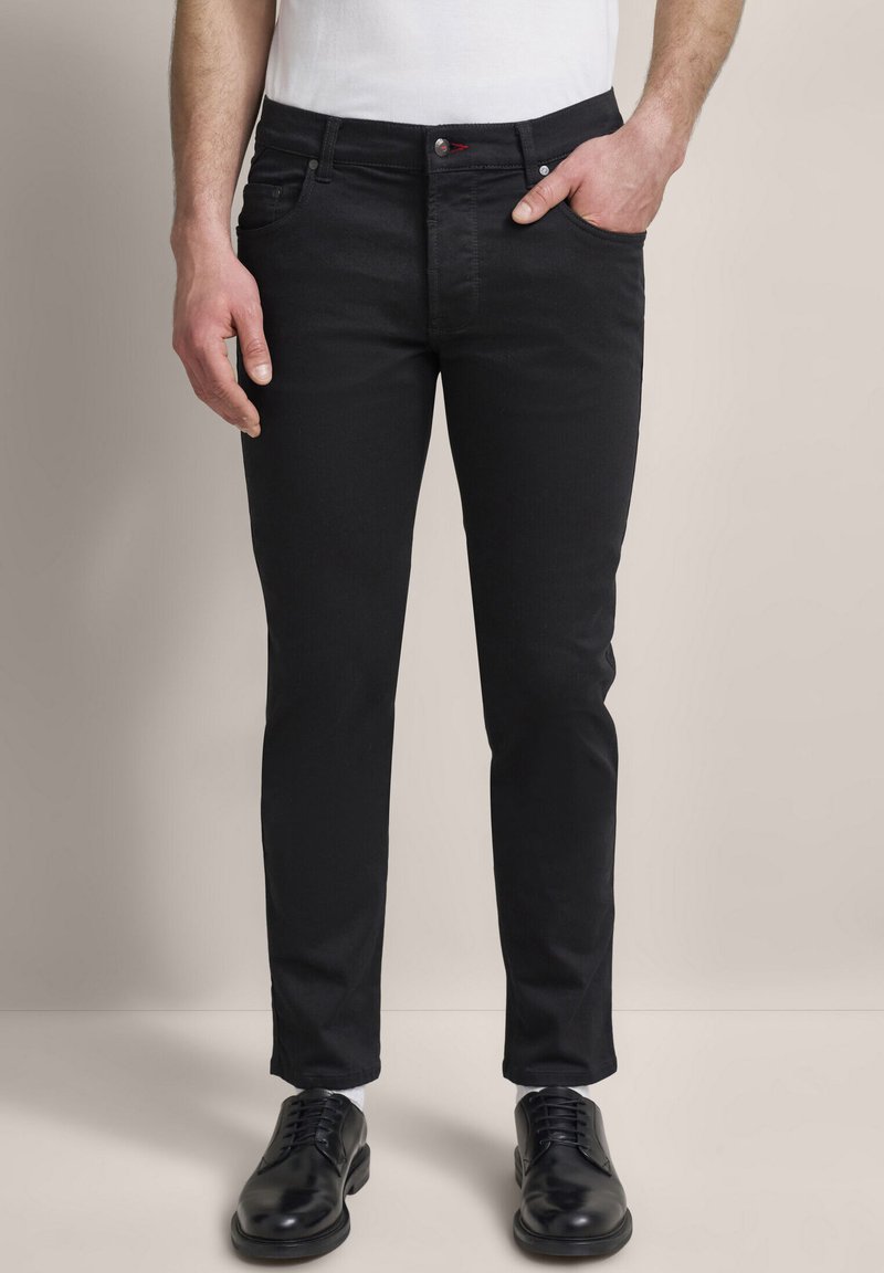Czarne dżinsy o slim fit, z klasycznym pięciopocketowym fasonem, czerwoną akcentującą guzik oraz gładką teksturą. W połączeniu z czarnymi butami.