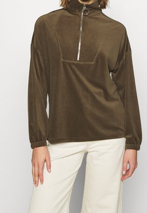 Personne portant un pull marron côtelé à manches longues avec une fermeture éclair devant et un pantalon clair, debout devant un fond uni.