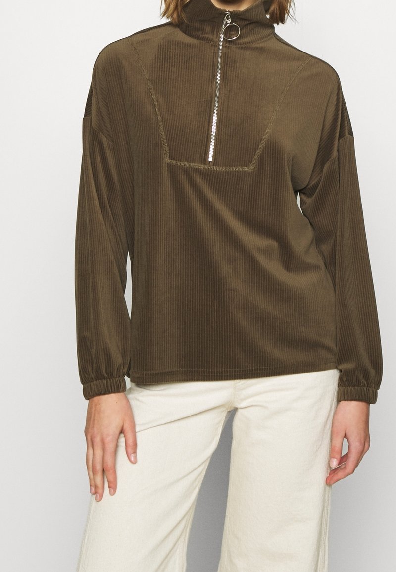 Personne portant un pull marron côtelé à manches longues avec une fermeture éclair devant et un pantalon clair, debout devant un fond uni.