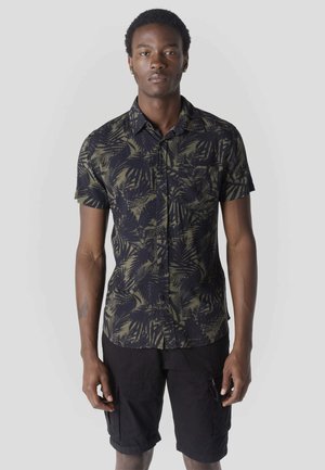 Joven con una camisa de manga corta abotonada con patrones de hojas en verde oscuro y negro, y pantalones cortos cargo negros, posando frente a un fondo liso.