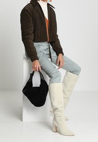 Giacca cropped in velluto a coste marrone con zip, top arancione, pantaloni azzurri chiari, stivali bianchi fino al ginocchio e borsa a tracolla nera.