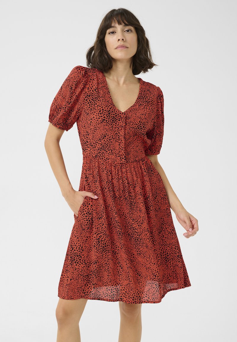 Robe rouge avec un motif à pois noirs, manches bouffantes courtes, décolleté en V et couture à la taille. Comprend deux poches latérales et un tissu léger et fluide.