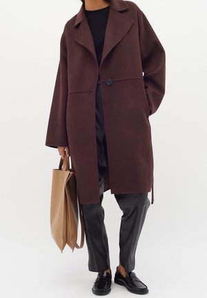 Osoba ubrana w ciemnobrązowy, oversize płaszcz, czarne spodnie, czarne mokasyny oraz niosąca dużą beżową torbę typu tote na jednolitym białym tle.