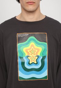 Čierne bavlnené tričko s mnohobarevným grafickým potlačou s vlnitými tvarmi a hviezdami. Potlač obsahuje zelené, žlté a modré odtiene.