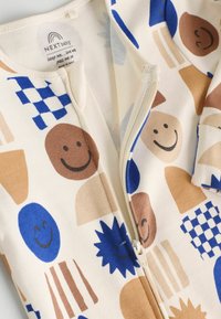 Mono de algodón con cremallera en color crema, que presenta patrones gráficos de caras sonrientes y formas geométricas en azul, marrón y beige. Textura suave.