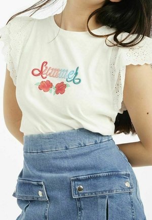 Vrouw die een wit shirt met gerimpelde mouwen en kleurrijke ""Summer"" tekst en bloemen draagt, gecombineerd met een hooggevoerde blauwe denim rok met zakken.
