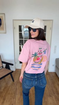 Mujer con camiseta rosa con diseño de tazón de frutas coloridas, jeans azules, gorra crema con cerezas y gafas de sol negras, sonriendo en interiores.