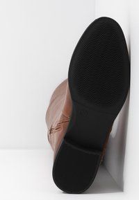 Botte en cuir marron avec une semelle en caoutchouc noire montrant une adhérence texturée, placée debout contre un fond d’angle blanc.