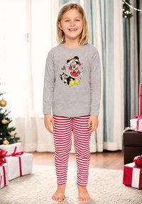 Una ragazza sorridente indossa una maglia grigia di Minnie Mouse per Natale e pigiama a righe rosse e bianche. Sta scalza in una stanza decorata con regali incartati.