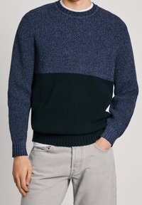 Mörkblå tröja i strukturerad stickning, med ett blockfärgat mönster av ljusare och mörkare nyanser. Ribbad ärm och nederkant. Bärs över en vit skjorta.