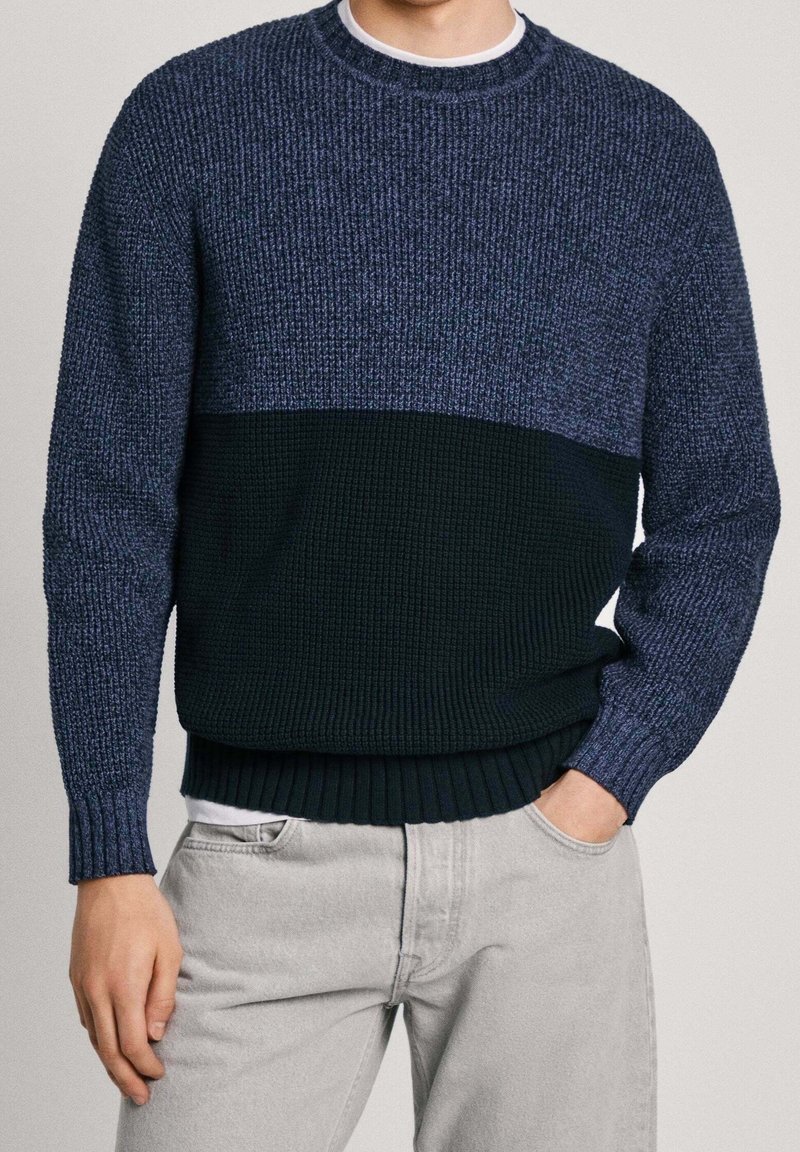 Mörkblå tröja i strukturerad stickning, med ett blockfärgat mönster av ljusare och mörkare nyanser. Ribbad ärm och nederkant. Bärs över en vit skjorta.