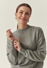 Pull gris à manches longues avec un col montant, texture douce, coupe décontractée et poignets côtelés ; porté les mains jointes près du col.