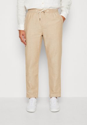 Beige Kordelhose mit elastischem Bund, kombiniert mit weißen Sneakern und einem weißen langärmeligen Hemd mit Knopfleiste.