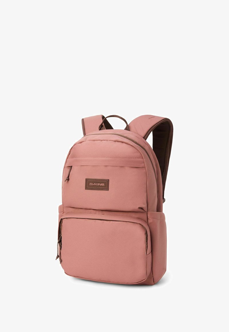 Sac à dos en tissu rose de forme rectangulaire, avec une poche avant, des sangles rembourrées et une poignée sur le dessus. Accents marron et détail du logo.