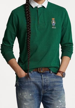 Person mit langem Zopf trägt grünes Langarm-Poloshirt mit Bären-Aufnäher, weißem Kragen, blauen Jeans mit Used-Look, braunem Gürtel und brauner Uhr.
