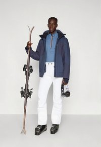 Síruha sötétkék dzsekivel, elöl cipzáras kék felsővel, fehér sínadrággal és fekete sícipővel. Síléceket és símaszkot tart a kezében.