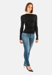 Donna con capelli castani lisci, che indossa una maglietta nera attillata a maniche lunghe, jeans skinny blu e tacchi alti neri, in piedi davanti a uno sfondo bianco.