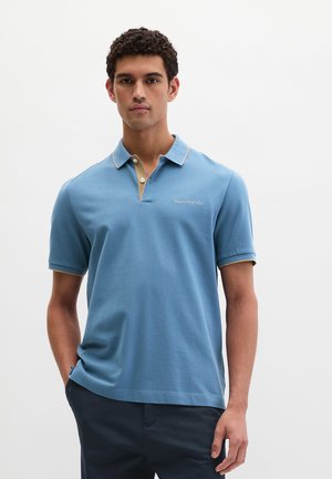 Jonge man draagt een lichtblauw poloshirt met korte mouwen, beige kraagafwerking en donkerblauwe broek, staand tegen een effen witte achtergrond.