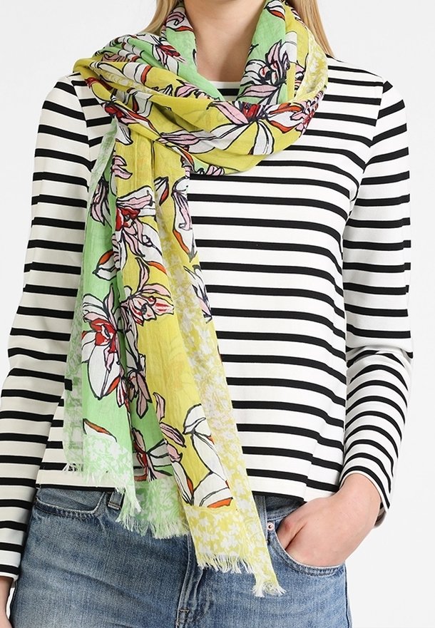 Femme portant un foulard jaune et vert à motifs floraux sur un t-shirt à manches longues rayé noir et blanc, avec un jean bleu et une main dans la poche.