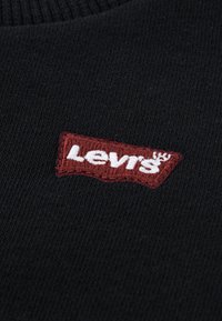 Černá svetrvová látka s červeně vyšívaným logem Levi's ve tvaru obdélníku na levé straně. Viditelný texturovaný pletený vzor.