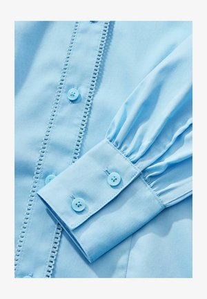 Blusa azzurro chiaro in tessuto morbido, con chiusura a bottone, dettagli sulla patta e maniche a sbuffo con polsini abbottonati.