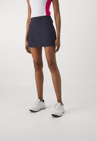 Kvinna klädd i en marinblå minikjol, vit och rosa ärmlös topp, och vita sneakers som står på en enkel ljusgrå golvyta.