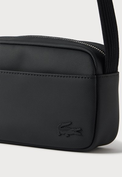 Крупен план на черна текстурирана чанта за рамо Lacoste с цип, регулируема презрамка и релефно лого с крокодил.