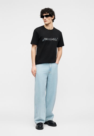 Νεαρός άνδρας που φοράει μαύρο γραφικό μπλουζάκι, ανοιχτό μπλε παντελόνι φαρδύ, μαύρα παπούτσια και μαύρα γυαλιά ηλίου, στέκεται με το ένα χέρι στην τσέπη.