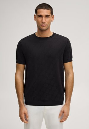 Mężczyzna ubrany w czarny sweter z krótkim rękawem i okrągłym dekoltem z subtelnym wzorem diamentów, w połączeniu z białymi spodniami na neutralnym tle.