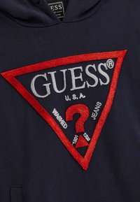 Felpa navy con un grande logo triangolare rosso e grigio con la scritta "GUESS" e "U.S.A.", dettagli di ricamo elaborati e un design di punto interrogativo.
