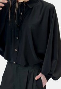Blusa nera con colletto e maniche lunghe e voluminose, caratterizzata da una chiusura frontale con bottoni e una texture liscia e lucida. Abbinata a pantaloni su misura.
