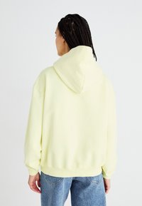 Felpa con cappuccio oversize di colore giallo chiaro, realizzata in tessuto morbido, dotata di cappuccio con cordino e polsini a costine, abbinata a jeans in denim blu.