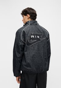 Mladi moški s kratkimi kodrastimi lasmi, oblečen v črno jakno Nike Air z vzorcem, obrnjeno v stran, ki prikazuje podroben logotip na hrbtni strani.