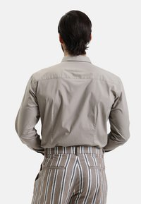 Camicia button-up grigia chiara con una texture liscia, dotata di yoke sul retro e colletto semplice, abbinata a pantaloni a righe marroni.