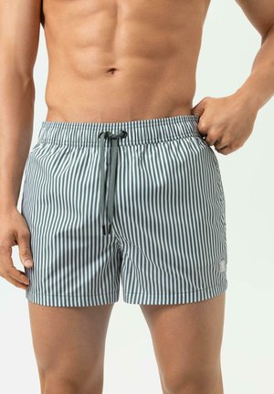 Man met groene en witte zwemshort met verticale strepen en trekkoord, met één hand zijn taille vasthoudend.