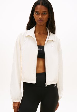 Mujer joven con cabello largo que lleva una chaqueta blanca con cremallera sobre un sujetador deportivo negro y leggings negros, de pie frente a un fondo blanco.