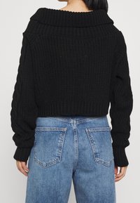 Black knitted sweater with a wide ribbed collar, cropped fit, and cable-knit pattern on sleeves, paired with light blue jeans.