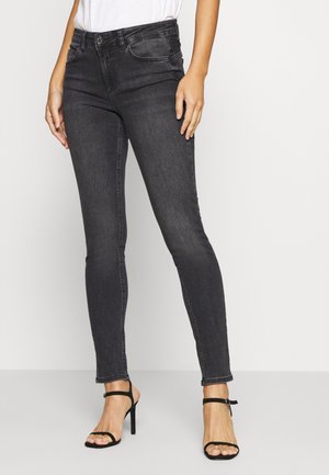 Vrouw die strakke zwarte skinny jeans en zwarte sandalen met hoge hakken draagt, staand tegen een effen witte achtergrond.