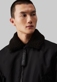Bomber negra con un cuello de borrego texturizado en marrón oscuro. Cuenta con un cierre de cremallera y una etiqueta con la marca "Calvin Klein". Tejido suave.