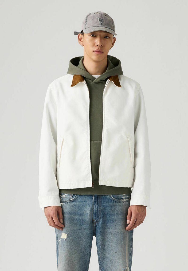 Chaqueta blanca con cuello de ante color tan, llevada sobre una sudadera con capucha verde. El modelo también lleva vaqueros azules desgastados y una gorra gris. Fondo neutro.