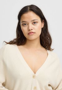 Beige stickad cardigan med djup V-ringning, som har tre stora knappar och textil med struktur. Designen har lösa, flödande ärmar.