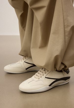 Weiße Schnürschuhe mit schwarzen Streifen, getragen zu locker sitzenden, beigen Hosen mit weitem Bein, stehend auf einem neutralen Boden.