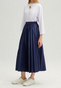 Camicia bianca, comoda, a maniche lunghe con scollatura tonda; gonna midi plissettata blu navy; sneakers bianche; collana con pendente dorato.