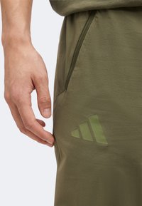 Gros plan d'une main à côté d'une poche de pantalon de sport vert olive avec un logo Adidas discret près de la cuisse.